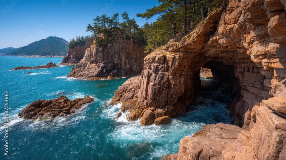 Obraz premium Rock formations rise majestically along a rugged coastline, with waves crashing against them. A natural cave is visible, surrounded by lush greenery against a clear blue sky