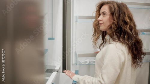 Woman carefully examining modern refrigerator in electronics store, slowly opening and closing doors and drawers while evaluating appliance features and design for potential purchase