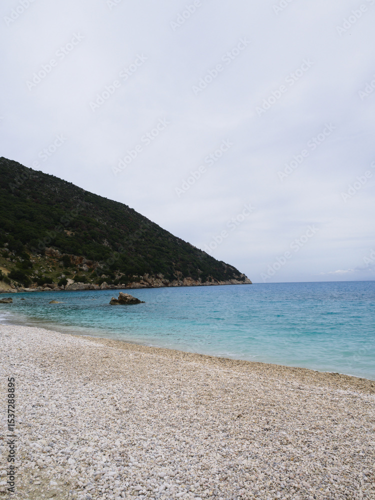 Fototapeta premium White gravel beach under cloudy sky in Kefalonia with clean coastline, perfect for commercial use and copyspace.