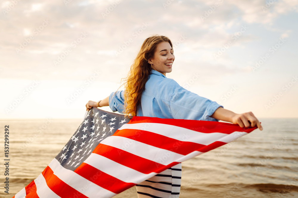 Fototapeta premium Happy woman on the beach with American flag on her shoulders. Beautiful woman enjoying sunset at sea, feeling freedom holding US flag. Independence and celebration concept.