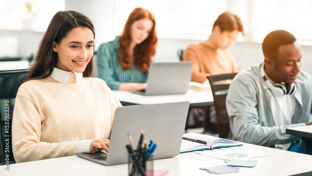 Modern Education, Technology And Lifestyle Concept. Group of diverse international students sitting at desk in classroom, selective focus on smiling female student using laptop, typing on keyboard Modern Education, Technology And Lifestyle Concept. Group of diverse international students sitting at desk in classroom, selective focus on smiling female student using laptop, typing on keyboard