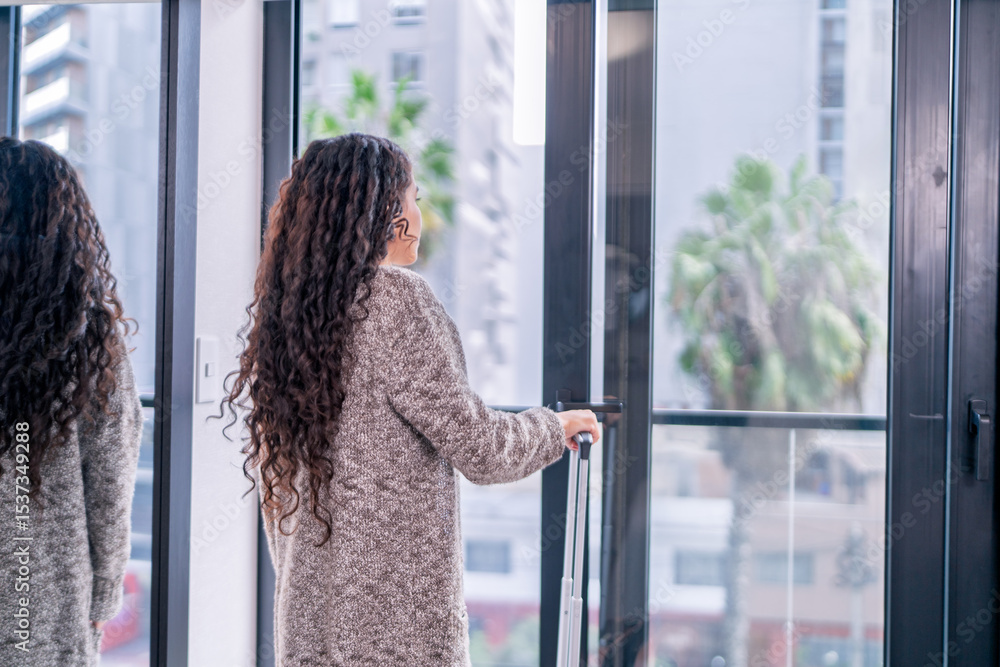 Obraz premium Digital nomad arriving at airbnb apartment with suitcase, gazing out window at city view