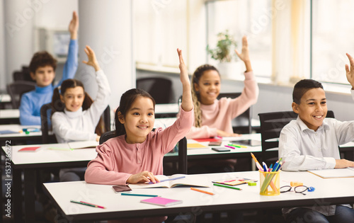 Return Back To School. Multiciltural diverse group of happy small elementary schoolchildren sitting at desks in classroom, raising hands up for an answer, learning with pleasure. Social distance