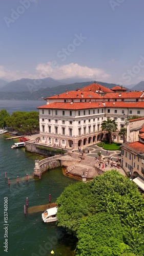 Drone video of Isola Bella’s Borromeo Palace seen from the lake. Elegant red-roofed estate rises above the water, surrounded by Alpine peaks, lush gardens, and sparkling Lake Maggiore, Italy