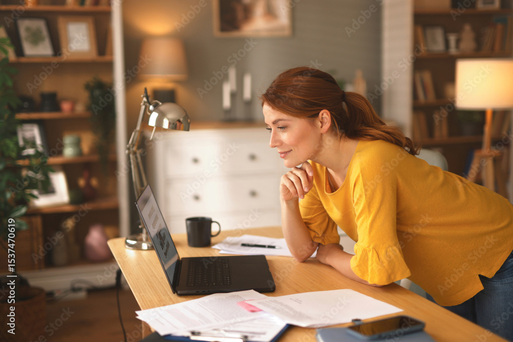 A woman in a yellow top leans over her laptop at a wooden desk in a bright living room, surrounded by papers and a coffee cup, focused on her work during the day. A woman in a yellow top leans over her laptop at a wooden desk in a bright living room, surrounded by papers and a coffee cup, focused on her work during the day.