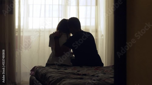 A couple sits together on a bed in a quiet room. The woman appears sad and emotionally exhausted. The man gently comforts her with a hug and kiss, offering support during a difficult time. 4K video