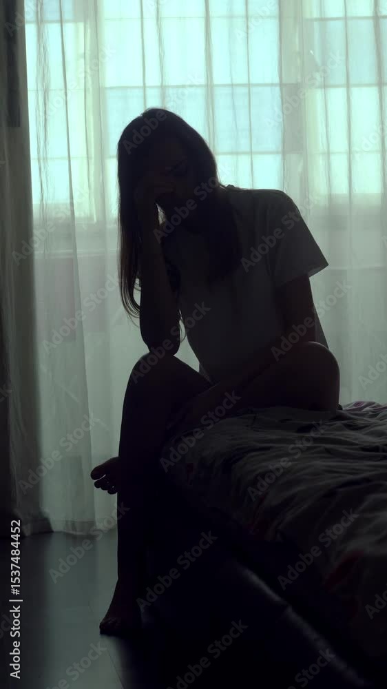 Vertical shot of a woman sitting alone on a bed in an empty room. Her silhouette reflects deep sadness, isolation, and emotional pain. A visual story of depression and mental struggle. Vertical shot of a woman sitting alone on a bed in an empty room. Her silhouette reflects deep sadness, isolation, and emotional pain. A visual story of depression and mental struggle.