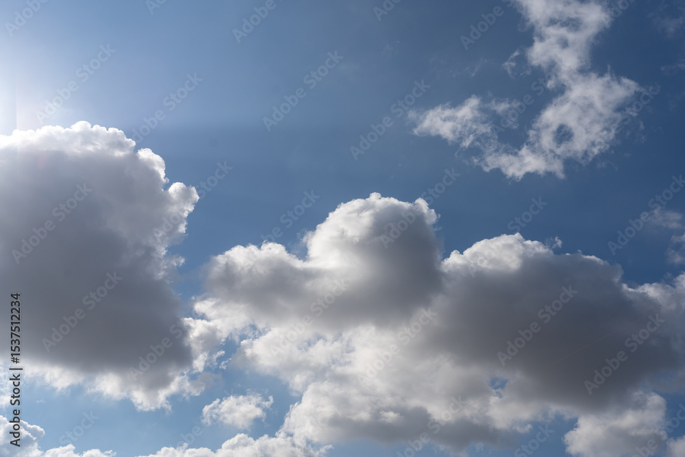 Fototapeta premium Fluffy white clouds drift across a vibrant blue sky under bright daylight. The sun shines brightly, creating a serene and inviting atmosphere. Perfect for a calm day outdoors.