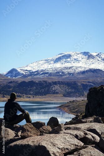 Konstfotografi Unrecognizable man sitting on rock looking at Lake Caviahue and snowcapped Copahue volcano in the background