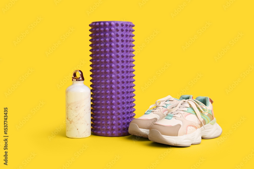 Fototapeta premium Foam roller with bottle of water and stylish sneakers on yellow background