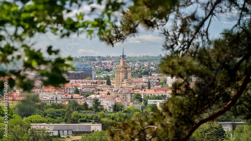 Central European city skyline framed by green trees. Features historic tower, modern buildings, residential areas under a partly cloudy sky. Ideal for travel & urban themes.