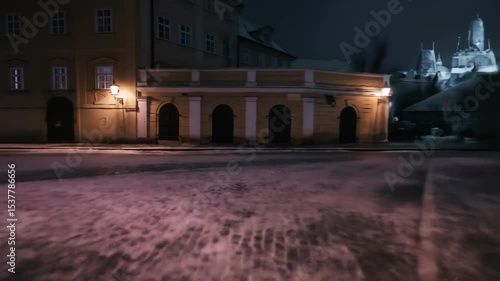 Low angle tracking shot over snowy cobblestone street at night. Illuminated by warm streetlights, the historic buildings and empty path evoke a quiet, mysterious European winter.