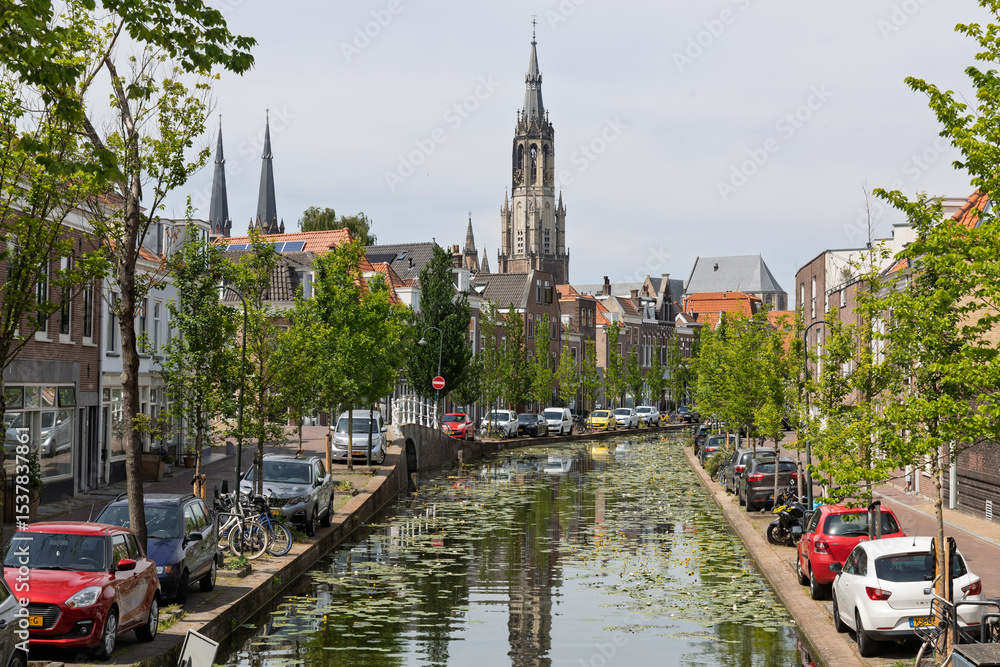 Naklejka premium A picturesque Delft canal view, featuring the majestic Nieuwe Kerk spire, charming canal-side houses, and parked cars under a sunny sky. Delft, Netherlands, 31 May 2025.
