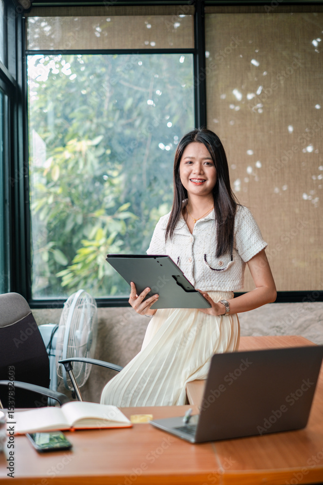 Confident young woman in a modern office setting, holding a tablet and smiling, with a laptop and large windows in the background. Confident young woman in a modern office setting, holding a tablet and smiling, with a laptop and large windows in the background.