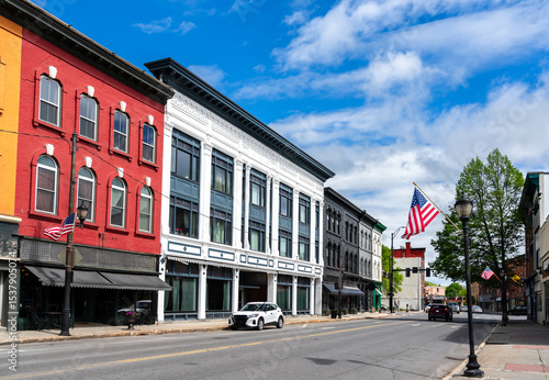 Fototapeta Naklejka Na Ścianę i Meble -  A row of red, white, and gray 19th-century storefronts lines Water Street in Augusta, Maine, United States, where American flags flutter beneath a bright spring sky.