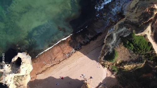 Hidden sandy beach with turquoise waters and waves crashing on the shore, surrounded by dramatic rocky cliffs and sea caves, captured from an aerial perspective.