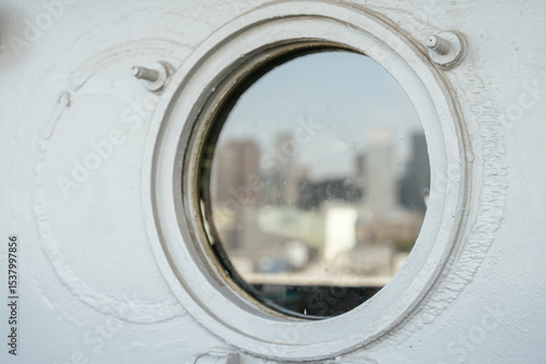 A circular porthole window on a white ship wall shows a soft, blurred view of a city skyline in the distance. The metal frame and bolts around the porthole are in sharp focus.