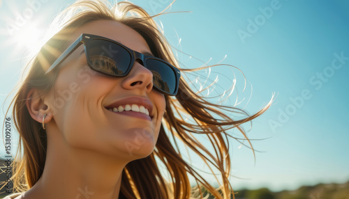 Confident woman wearing sunglasses smiles outdoors with sunlight and blue sky, enjoying bright and windy day