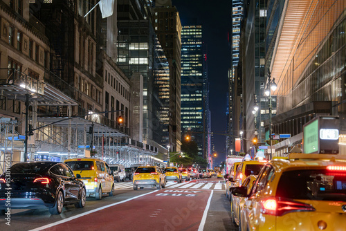 Photography Yellow taxis and a black car drive through Midtown Manhattan at night under skyscrapers and streetlights