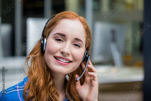 Foto Red-haired support rep using headset and blue plaid shirt smiling at open-plan o