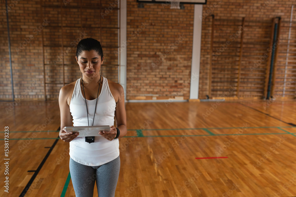 Fototapeta premium Athletic coach is holding tablet and wearing whistle on wooden basketball court under hoop