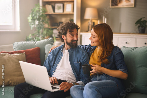 Photography Two people are sitting closely on a sofa in a cozy living room, engaging with a laptop and sharing a warm moment together with smiles and conversation