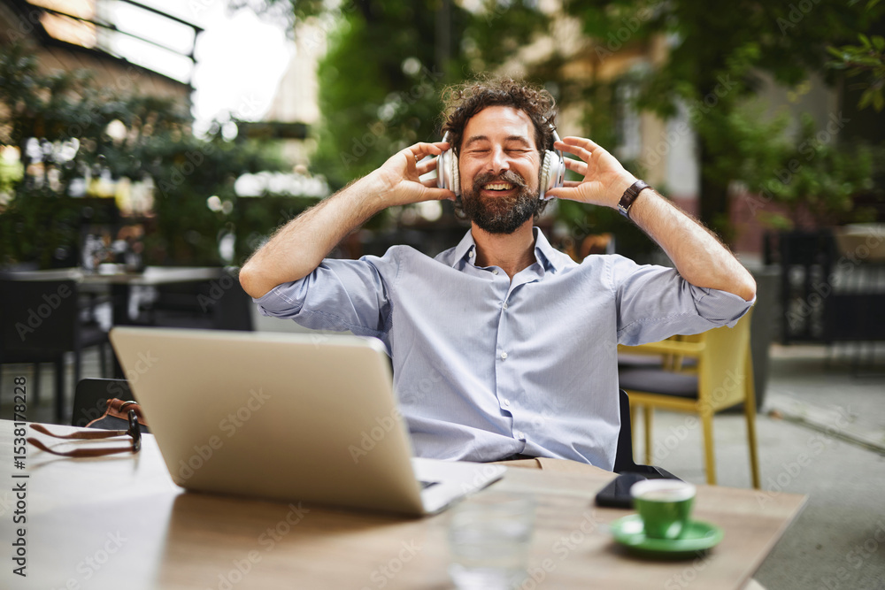A man relishes a moment of relaxation as he listens to music through headphones while working on a laptop at an outdoor cafe. Sunlight brightens the urban setting filled with greenery. A man relishes a moment of relaxation as he listens to music through headphones while working on a laptop at an outdoor cafe. Sunlight brightens the urban setting filled with greenery.