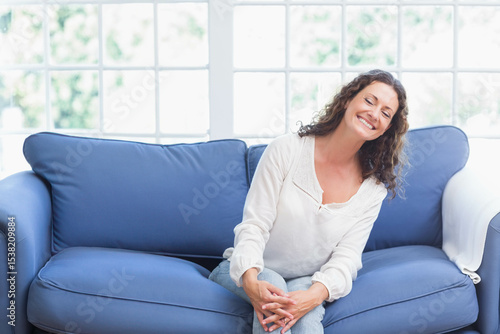 Free Download Mid adult woman sitting on blue sofa by multi-pane window in bright room, smiling, copy space - GFXLoad