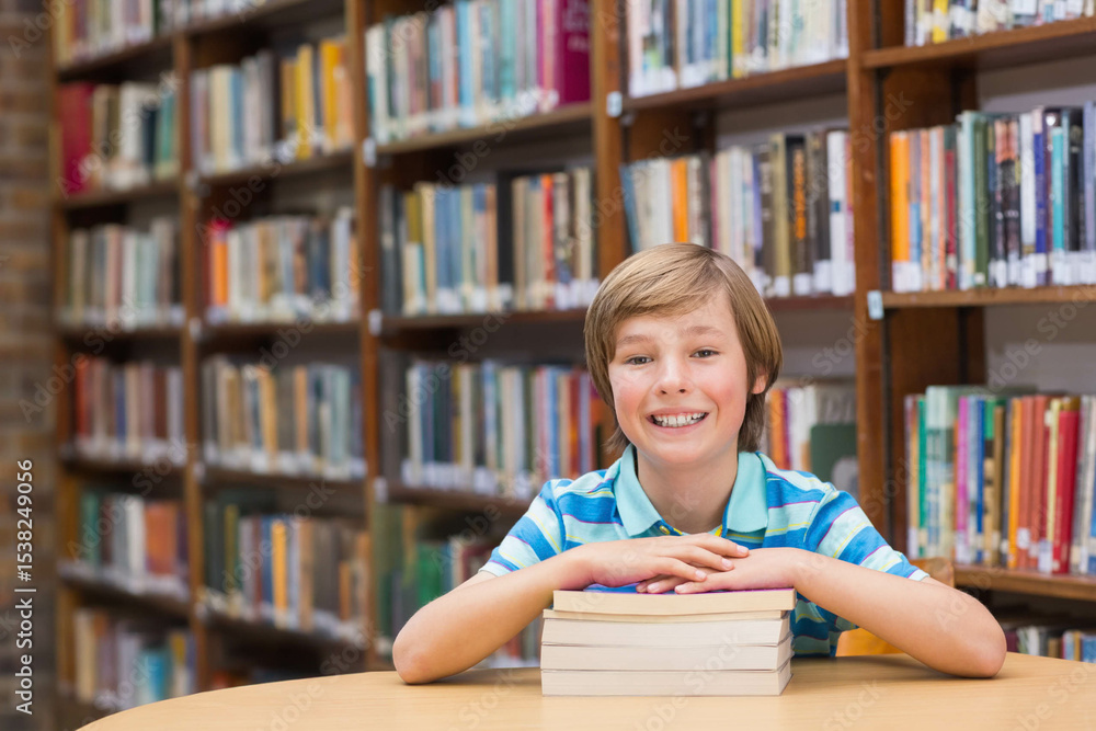 School-aged boy sitting at round table in library resting on three hardcovers with bookshelves School-aged boy sitting at round table in library resting on three hardcovers with bookshelves
