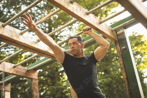 Man gripping wooden monkey bars and reaching next rung in leafy park with dappled sunlight
