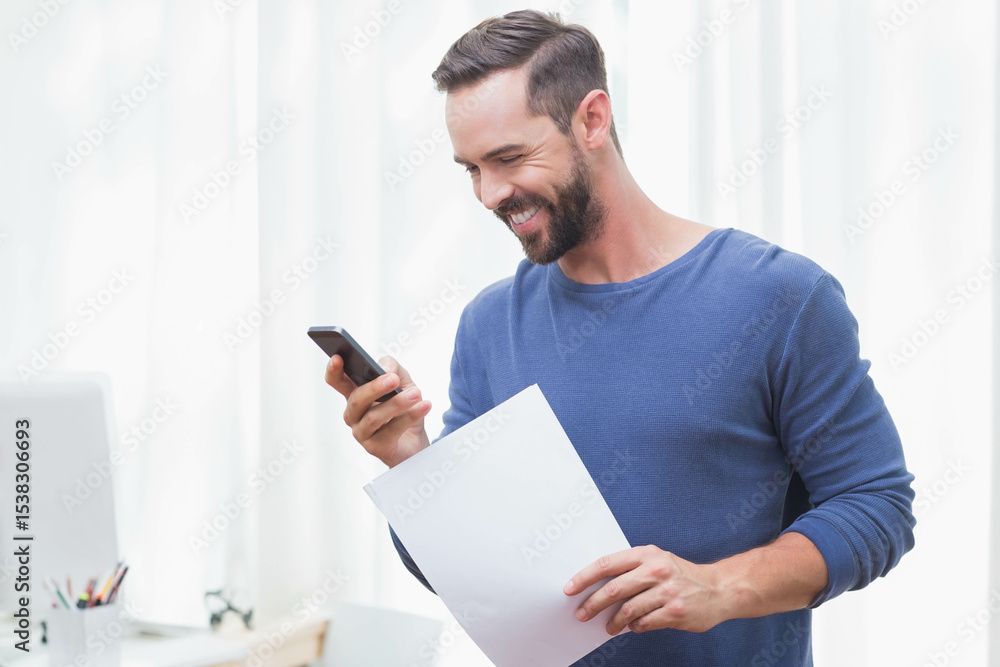 Man in mid-30s standing in home office holding blank paper checking smartphone with laptop visible Man in mid-30s standing in home office holding blank paper checking smartphone with laptop visible