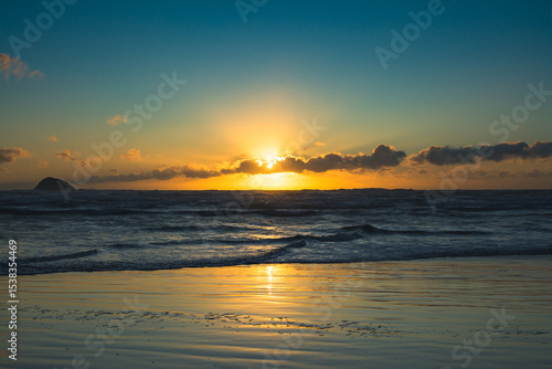 The sun sets behind a veil of clouds over Muriwai Beach, casting golden reflections across the rippling tide and wet sand in a tranquil, breathtaking coastal scene