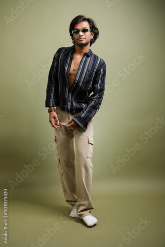 Stylish young man posing in striped shirt and cargo pants with accessories on green background.