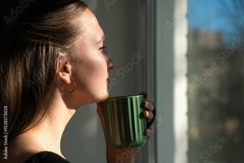 Young woman enjoys a quiet moment, sipping coffee and gazing out the window at the sunny cityscape, lost in thought and finding solace in the simple pleasures of life