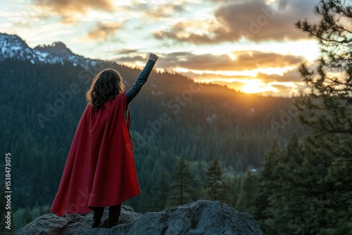 Young girl dressed as a superhero raises her arm triumphantly at sunset atop a rocky outcrop surrounded by mountains and trees
