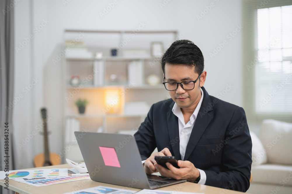 Professional man in suit working on laptop and texting on smartphone in modern office with charts and documents on table, business concept, technology use Professional man in suit working on laptop and texting on smartphone in modern office with charts and documents on table, business concept, technology use