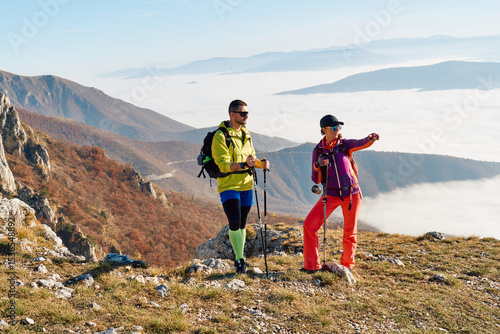 Couple hikers trek up a rocky mountain trail, enjoying a scenic view above the clouds, with majestic peaks in the background.