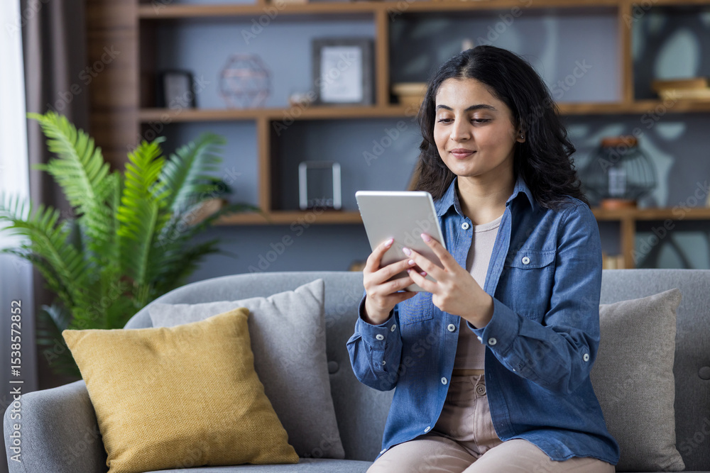 Young Indian girl sitting on sofa at home, using tablet, talking on video call Young Indian girl sitting on sofa at home, using tablet, talking on video call