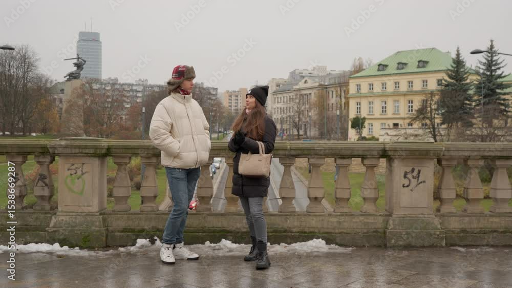 A young couple stands on a historic bridge in Warsaw, Poland, on a cold winter day. They share a warm, friendly conversation, enjoying the urban cityscape and their European travel adventure together. A young couple stands on a historic bridge in Warsaw, Poland, on a cold winter day. They share a warm, friendly conversation, enjoying the urban cityscape and their European travel adventure together.