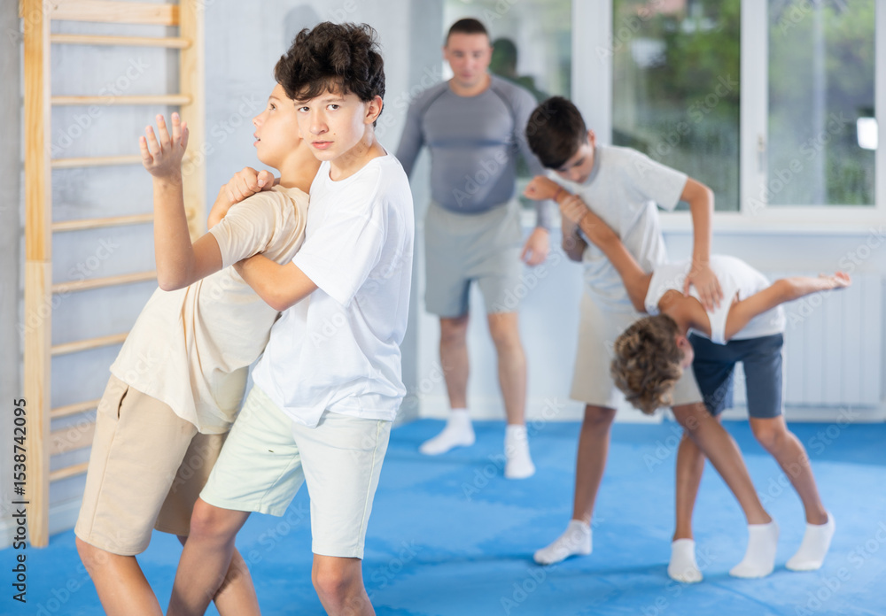 Naklejka premium Focused teen boy practicing basic protection skills with opponent in training bout during self defense course with coach in gym, trying to escape rear bear hug technique..