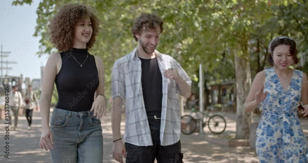 Three friends enjoy a sunny day, laughing and talking as they walk together along a paved path lined with trees. Three friends enjoy a sunny day, laughing and talking as they walk together along a paved path lined with trees.