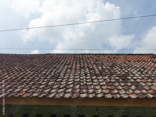 Close-up view of an old, weathered terracotta tiled roof under a bright sky with soft clouds and power lines.