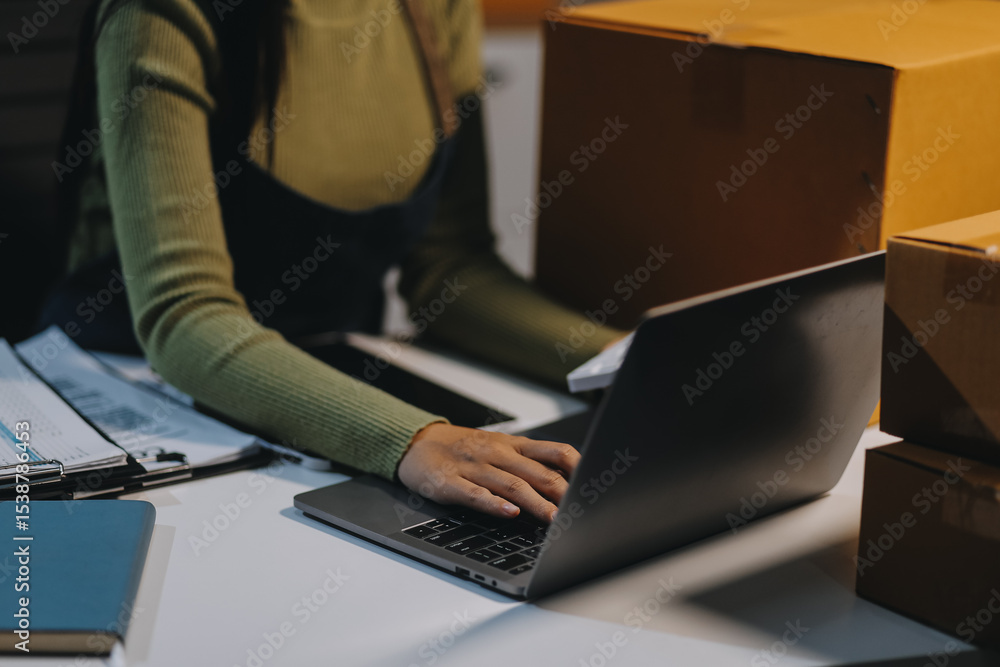 Fototapeta premium Startup SME small business entrepreneur of freelance Asian woman wearing apron using laptop and box to receive and review orders online to prepare to pack sell to customers, online sme business ideas.