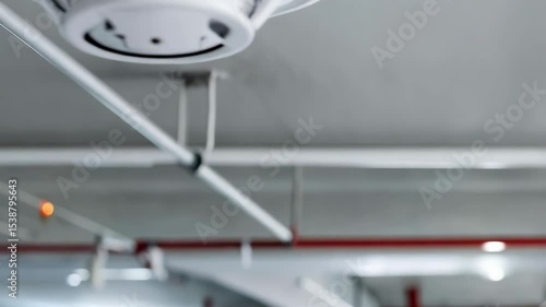 Indoor view featuring a white smoke detector mounted on ceiling with visible pipes, wires and overhead lighting system for safety monitoring.