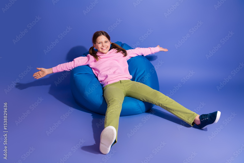 Obraz premium Adorable happy schoolgirl sitting on a comfortable blue bean bag against a vibrant purple background