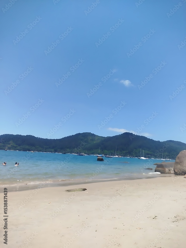 Fototapeta premium View of a sandy Ubatuba beach with mountains in the background and boats on the water under a clear blue sky