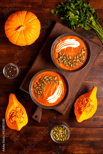 Top-down image of two bowls of pumpkin soup on a wooden board, decorated with cream swirls and pumpkin seeds, surrounded by fresh parsley, whole and halved pumpkins, and black pepper. Proper nutrition