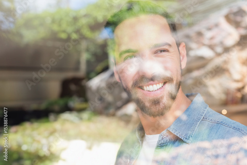 Mid adult male wearing white t-shirt and denim shirt standing inside home behind glass pane smiling