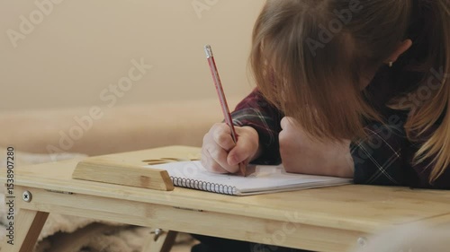 Young girl drawing on a notebook at home. Red-haired girl drawing and writing on a notebook placed on a small wooden table, in various moments of concentration