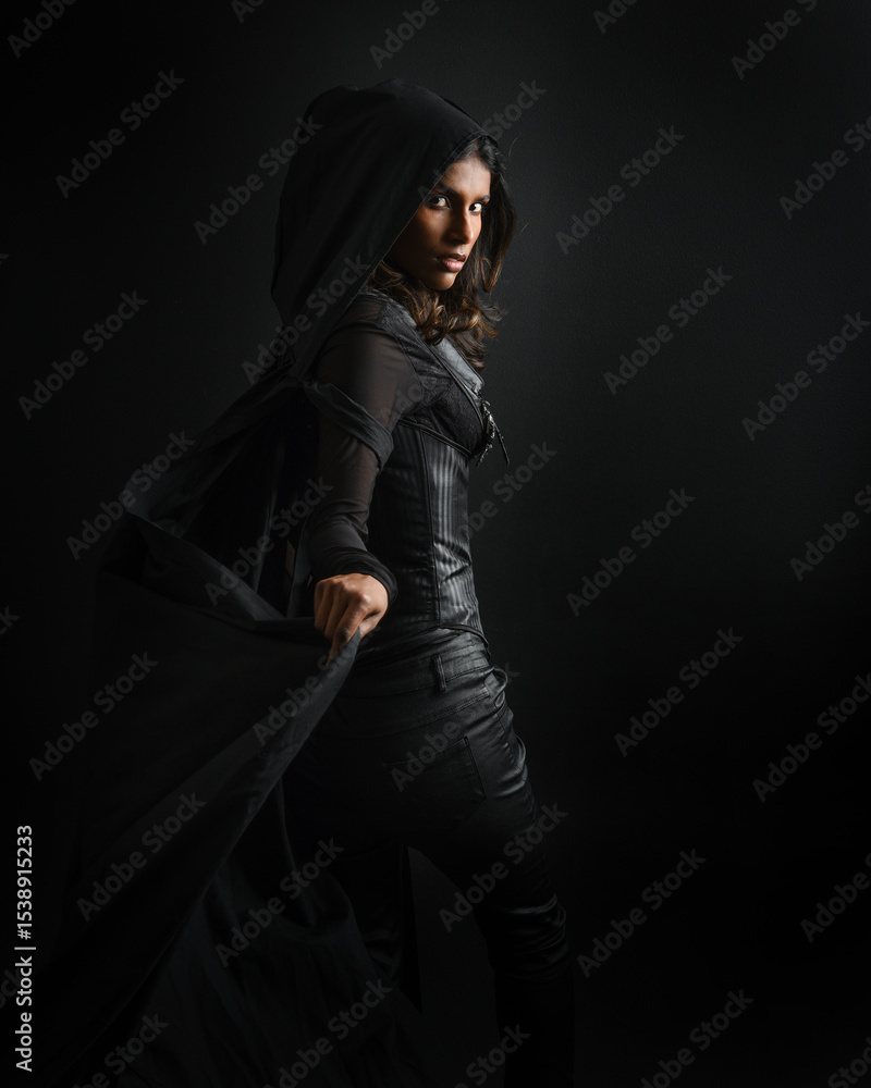 Naklejka premium portrait of mysterious hooded woman in black cloak, gothic fantasy or dystopian character. posing with gestural magical hand poses. lit with moody shadows, studio scene on dark isolated background